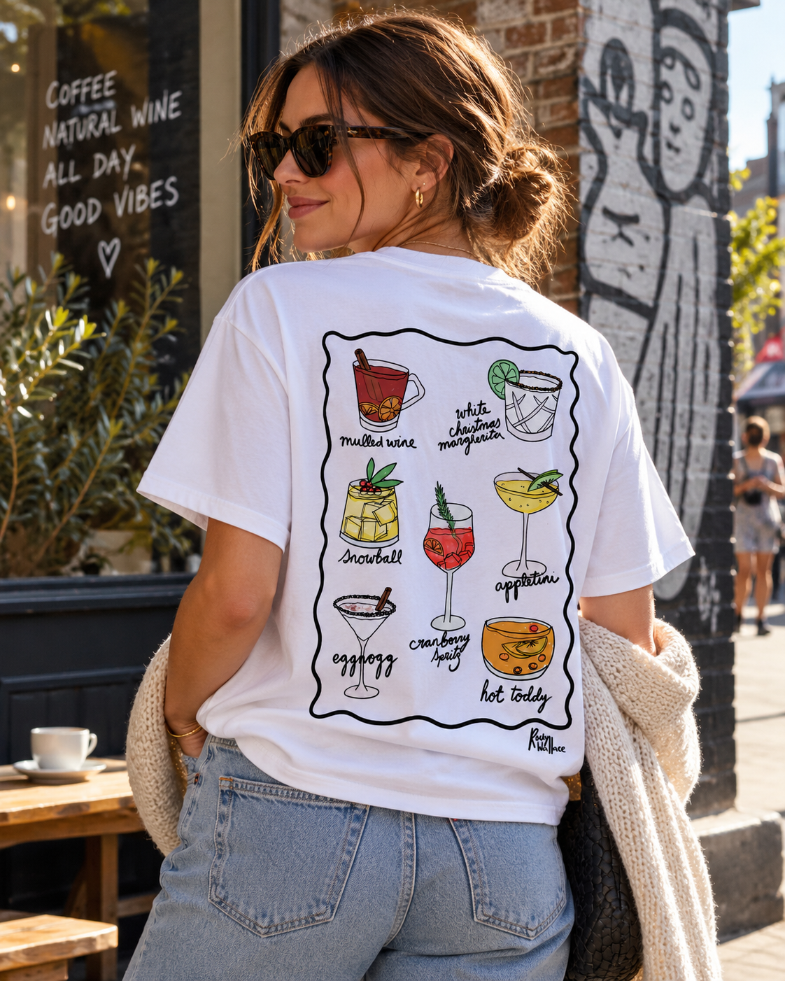 A woman wearing a white graphic t-shirt with a colorful illustration of Christmas cocktails on the back, paired with light blue jeans and a cream knit cardigan, photographed outdoors on an East London street outside a cafe