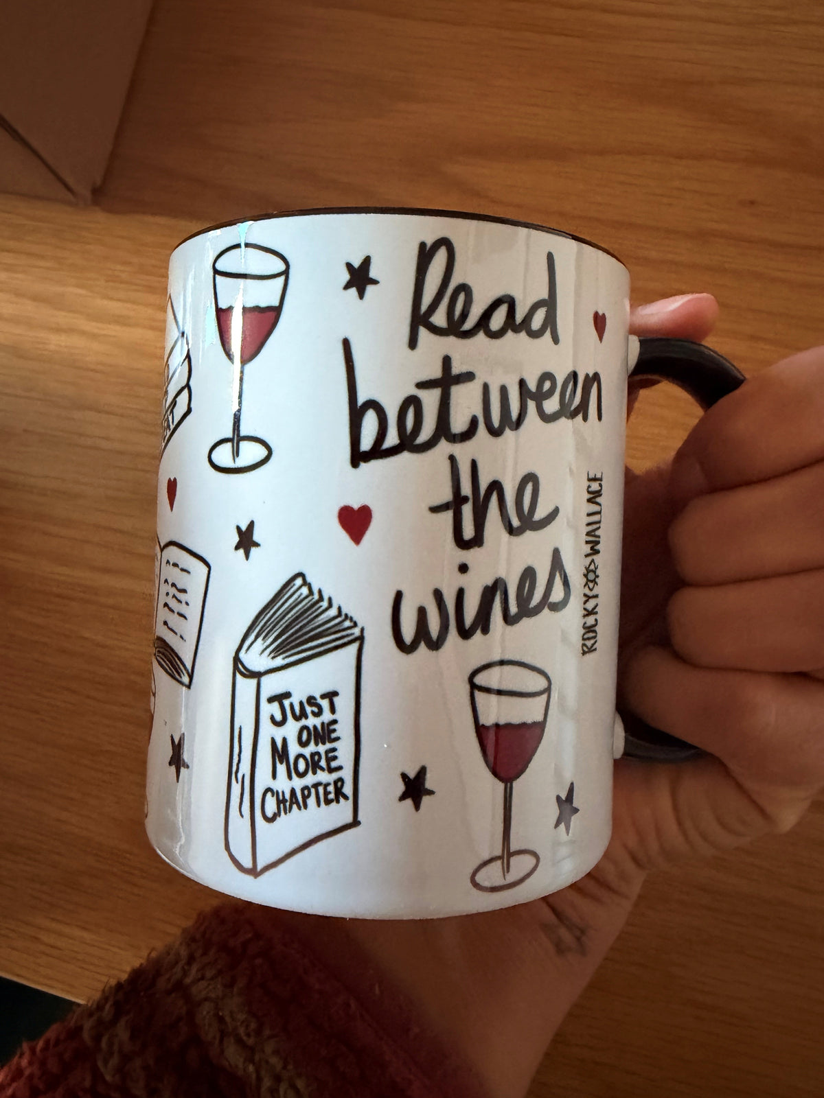 A hand holding a white ceramic mug with a black handle, decorated with illustrations of wine glasses, stars, and books, alongside the text 'Read between wines' and 'Just one more chapter'. The mug is photographed on a wooden table.
