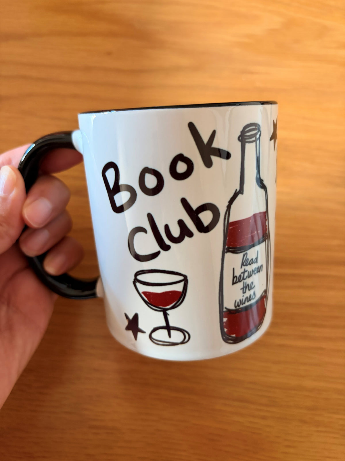 A hand holding a white ceramic mug with a black handle and black interior, printed with the words 'Book Club' and illustrations of a wine glass and a wine bottle on a wooden tabletop.
