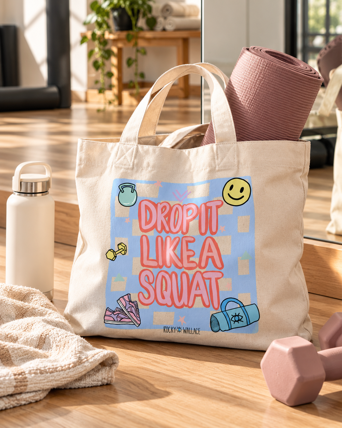 A beige canvas tote bag with a colorful graphic that reads 'DROP IT LIKE A SQUAT' in pink letters on a light blue patterned background. The bag is styled on a wooden floor with a pink yoga mat, a white water bottle, a towel, and a pink dumbbell.