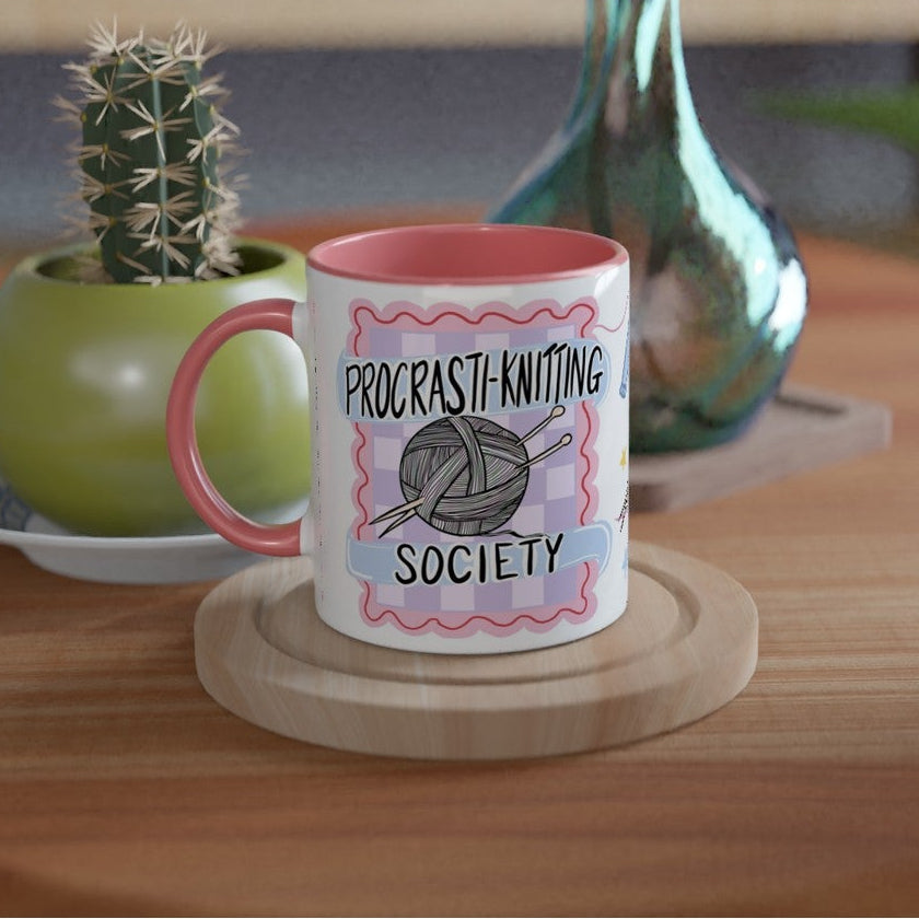 A white ceramic mug with a pink handle and interior, printed with the text 'PROCRASTI-KNITTING SOCIETY' and a knitting ball graphic, sitting on a round wooden coaster on a wooden table.