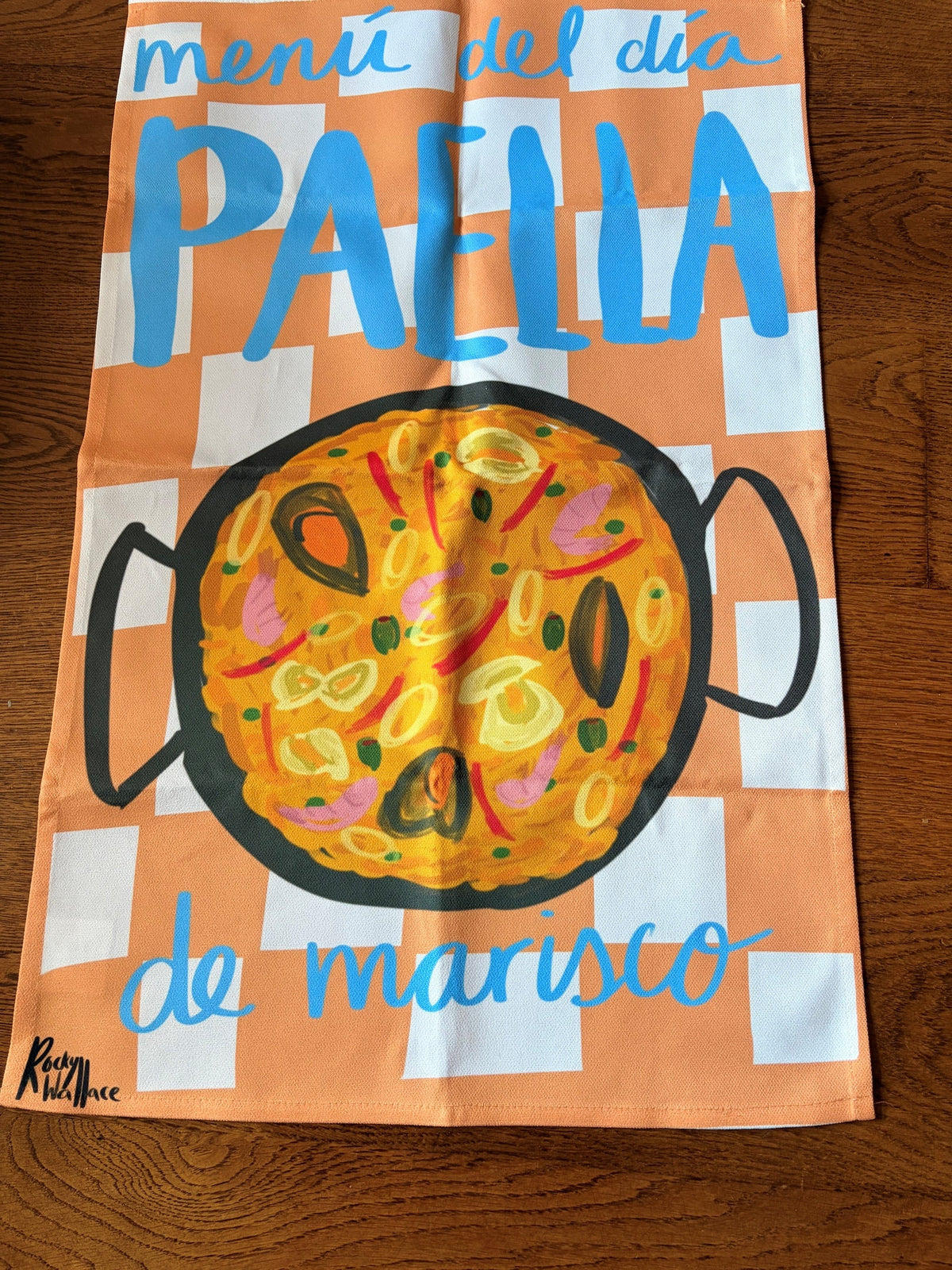 A rectangular kitchen towel with a hand-painted style illustration of a paella pan with paella, set against an orange and white checkered background with the text 'menú del día PAELLA de marisco'. The towel is laid flat on a dark wood surface.