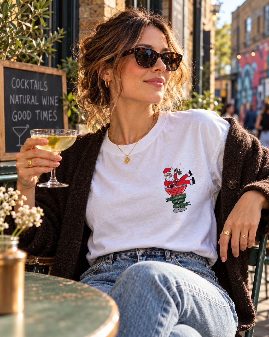 A woman sitting at an East London outdoor cafe wearing a white graphic t-shirt with a Christmas Santa design, a brown cardigan, and blue jeans, holding a cocktail in a glass.