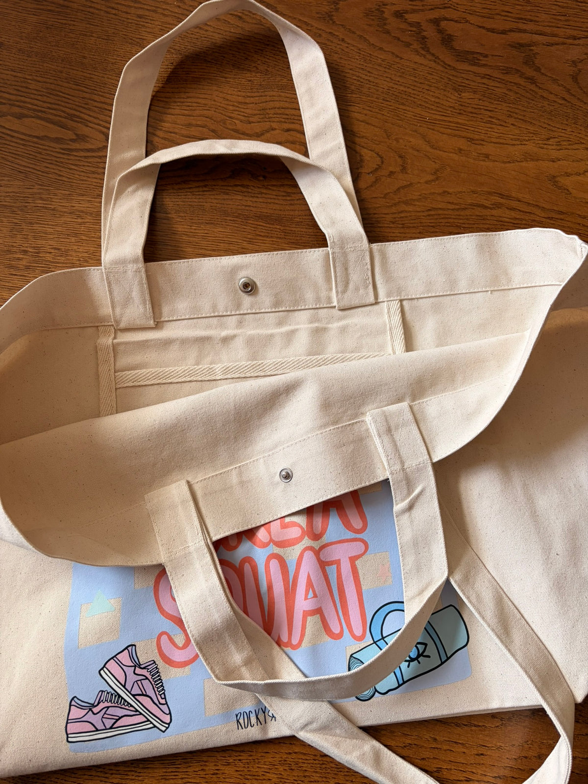 A natural beige canvas tote bag with metal snap buttons and long handles, laid flat on a brown gym studio floor, featuring snap closure and bright gym print on the front